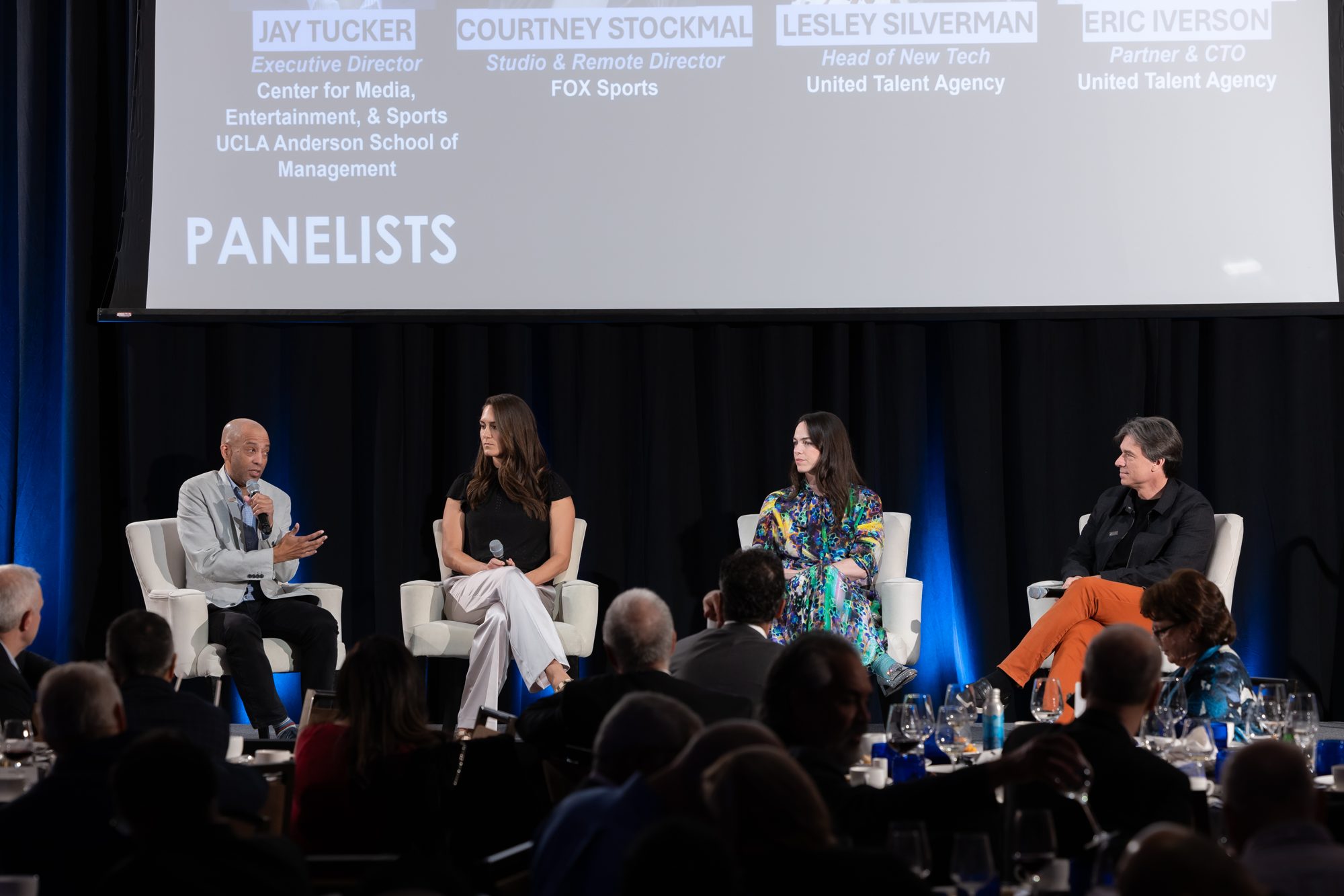 A panel discussion on stage at a leadership conference, with attendees seated at dinner tables
