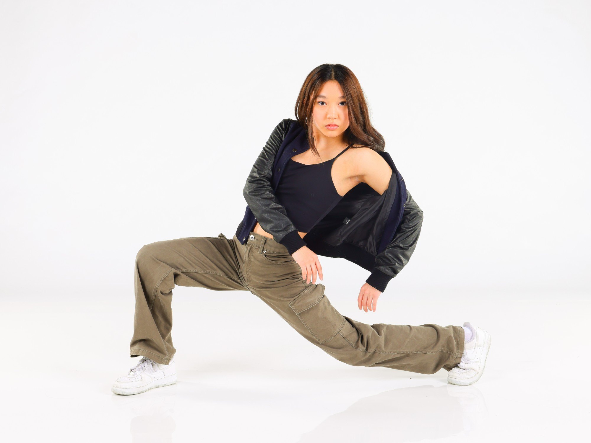 A hip hop dancer in a varsity jacket and cargo pants in a deep lunge on a white set