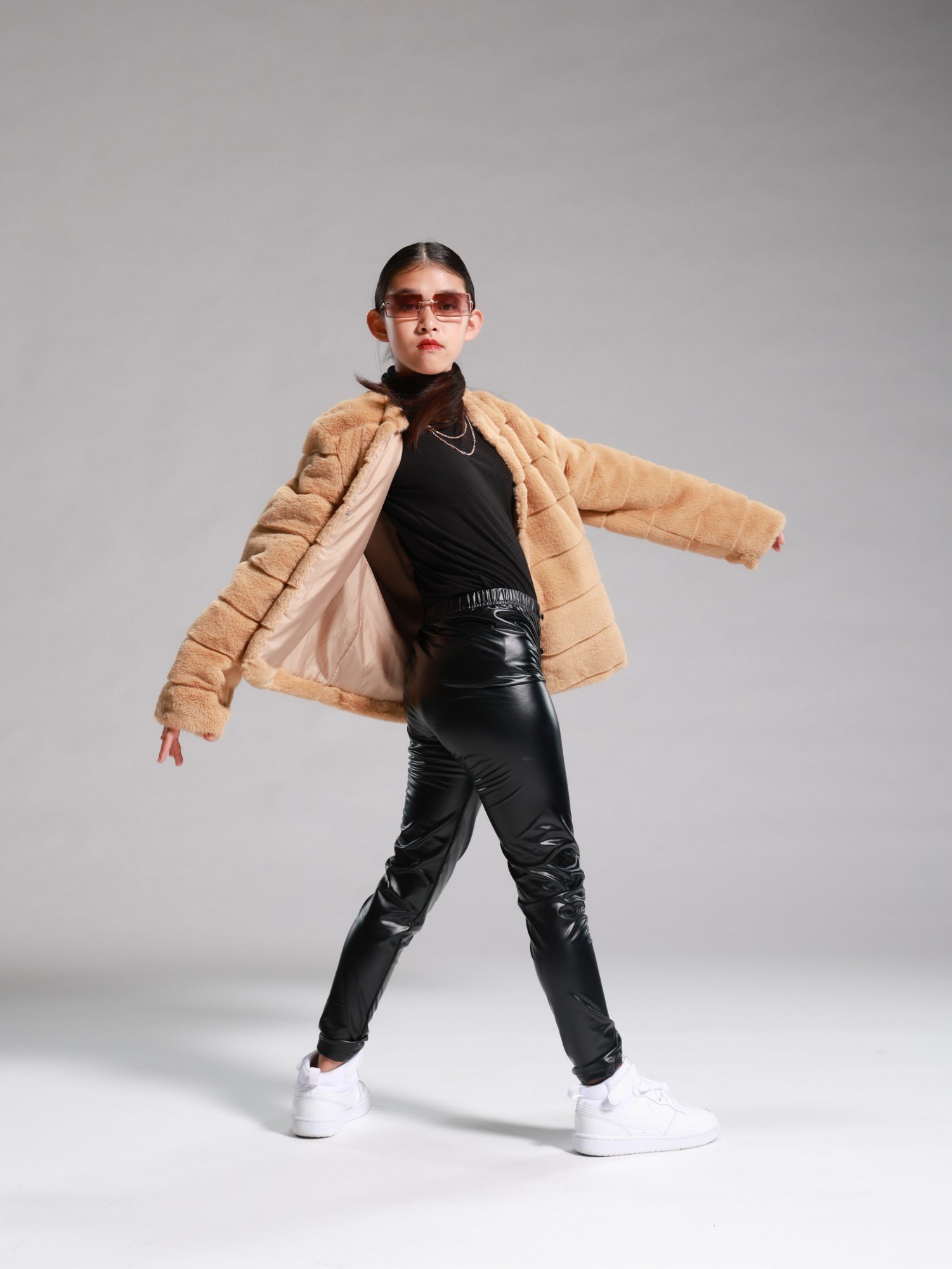 A young dancer in sunglasses and a faux fur jacket striking a pose on a grey backdrop